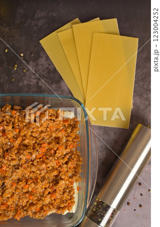 Top view. Bolognese sauce in a glass form. Lasagne pasta sheets, pepper mill. Black background. Homemade lasagna cooking preparation 123004252