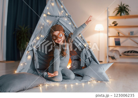 In pilot glasses. Cute little girl playing in the tent that is in the domestic room In pilot glasses. Cute little girl playing in the tent that is in the domestic room 123006235