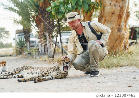 Male tourist in casual clothes and hat sitting and petting a cheetah outdoors Male tourist in casual clothes and hat sitting and petting a cheetah outdoors 123007463