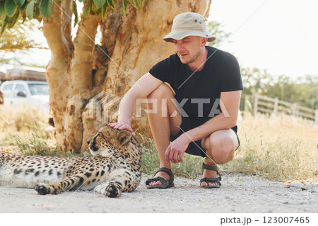 Male tourist in casual clothes and hat sitting and petting a cheetah outdoors 123007465