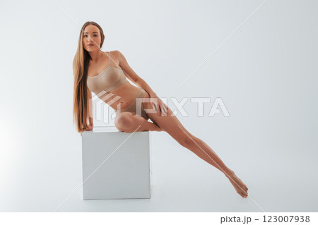 Sitting on the one leg on the white cube. Woman in underwear with slim body type is posing in the studio Sitting on the one leg on the white cube. Woman in underwear with slim body type is posing in the studio 123007938