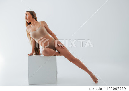 Sitting on the one leg on the white cube. Woman in underwear with slim body type is posing in the studio Sitting on the one leg on the white cube. Woman in underwear with slim body type is posing in the studio 123007939