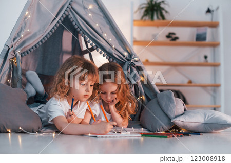 Learning to write and draw. With paper and pencils. Two little girls is in the tent in domestic room together 123008918
