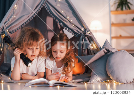 Reading book and using flashlight. Two little girls is in the tent in domestic room together 123008924