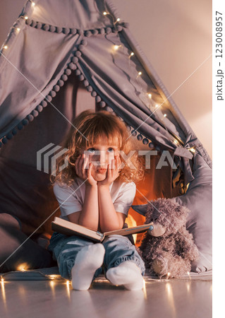 Sitting with book. Cute little girl with curly hair is in the tent in domestic room Sitting with book. Cute little girl with curly hair is in the tent in domestic room 123008957