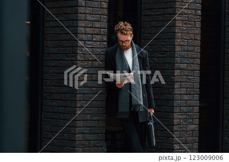 With newspaper in hands. Stylish man with beard and in glasses is outdoors near building 123009006