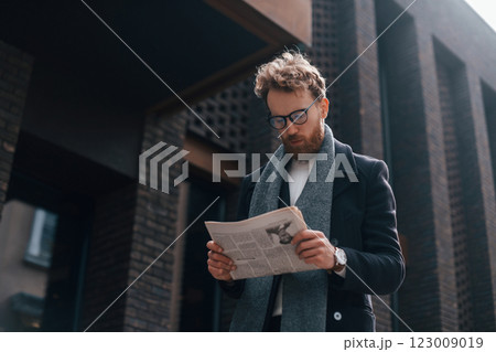 With newspaper in hands. Stylish man with beard and in glasses is outdoors near building With newspaper in hands. Stylish man with beard and in glasses is outdoors near building 123009019
