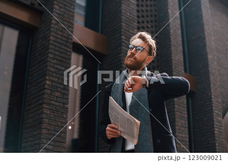 Checking time. Stylish man with beard and in glasses is outdoors near building 123009021