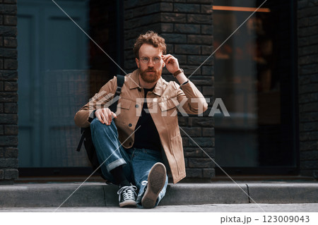 Sitting on the sidewalk. Stylish man with beard in khaki colored jacket and in jeans is outdoors near building Sitting on the sidewalk. Stylish man with beard in khaki colored jacket and in jeans is outdoors near building 123009043