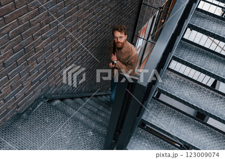 Looking up. Man in khaki colored jacket and in jeans is on the stairs on the building outdoors 123009074