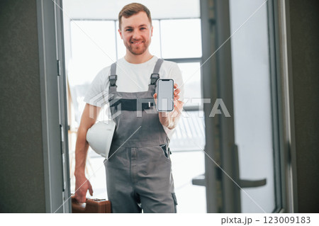 With smartphone in hand. Repairman is standing indoors in the modern room With smartphone in hand. Repairman is standing indoors in the modern room 123009183