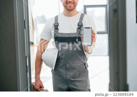 With smartphone in hand. Repairman is standing indoors in the modern room 123009184