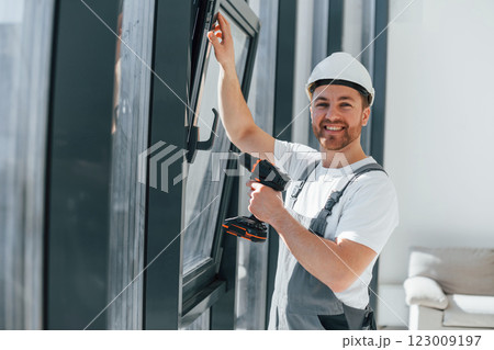 Using drill by installing windows. Repairman is working indoors in the modern room Using drill by installing windows. Repairman is working indoors in the modern room 123009197