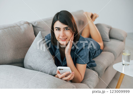 Holding phone and laying down on the sofa. Young woman is at home. Conception of beauty 123009999