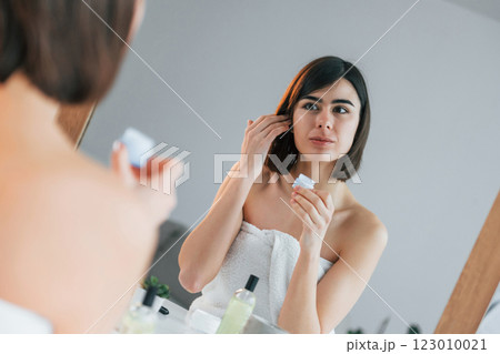 Sitting by the mirror and using cream on face. Young woman is at home. Conception of beauty 123010021