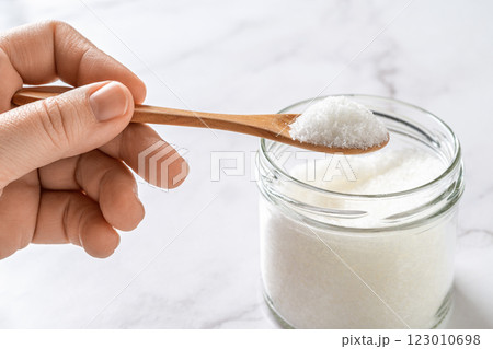 Woman fingers hold wooden spoon full of monosodium glutamate white crystals over glass jar. 123010698