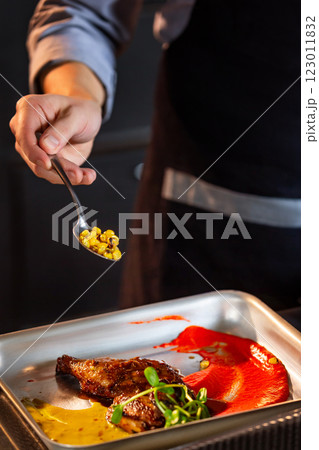 Chief cook is decorating a meat dish with corn at kitchen background. Vertical image. Chief cook is decorating a meat dish with corn at kitchen background. Vertical image. 123011832