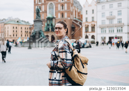 Tourist happy woman in glasses with backpack on Market Square in Krakow, Traveling Europe in autumn. St. Marys Basilica, cloudy day. Attractive female is exploring city. Top tourist attraction 123013504