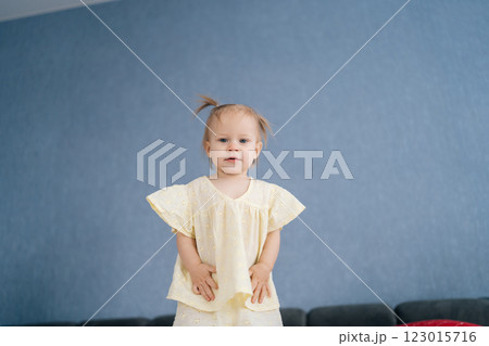 Portrait of pretty toddler girl wearing light yellow dress, standing on bed against blue wall, smiling looking at camera. Concept of simple charming scene of childhood innocence. Portrait of pretty toddler girl wearing light yellow dress, standing on bed against blue wall, smiling looking at camera. Concept of simple charming scene of childhood innocence. 123015716