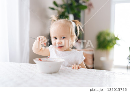 Portrait of cute toddler girl happily eating healthy beet borscht soup with spoon, savoring meal at home, surrounded by warmth and love in cozy kitchen room with bright interior. Portrait of cute toddler girl happily eating healthy beet borscht soup with spoon, savoring meal at home, surrounded by warmth and love in cozy kitchen room with bright interior. 123015736