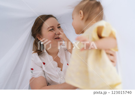 Cheerful young mother and infant daughter enjoying playful moments together, hiding under soft white blanket, pointing fingers and sharing laughter at home. Concept of parenting love and care. 123015740