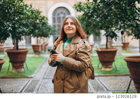 Happy 30s redhead woman using phone walking in city street in old town. Lifestyle, travel, tourism, technology, blogging, communication concept. Buildings with potted plants. Charming facade Happy 30s redhead woman using phone walking in city street in old town. Lifestyle, travel, tourism, technology, blogging, communication concept. Buildings with potted plants. Charming facade 123016129