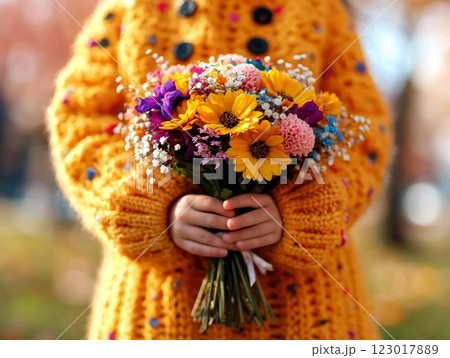 child in bright yellow sweater joyfully holds bouquet of colorful wildflowers while walking in spring parade. Blooming trees create soft backdrop of natural beauty. 123017889