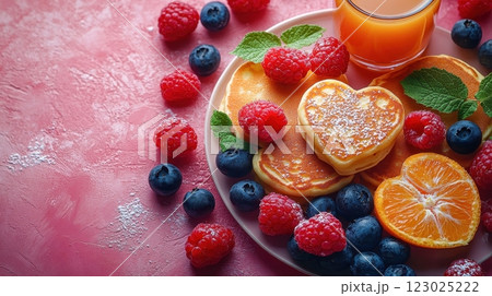 A romantic breakfast featuring fluffy pancakes topped with heart-shaped jam and surrounded by vibrant raspberries, blackberries, and blueberries, paired with a refreshing glass of orange juice. 123025222