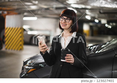 Confident Caucasian woman in business attire holding coffee and smartphone. Inside parking garage, she smiles while texting or checking messages. Outdoor work environment, professional engagement. Confident Caucasian woman in business attire holding coffee and smartphone. Inside parking garage, she smiles while texting or checking messages. Outdoor work environment, professional engagement. 123027401
