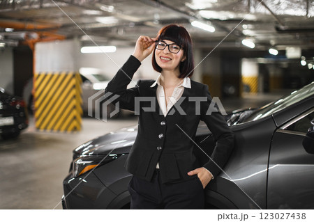 Young adult Caucasian female in business attire standing confidently near car in parking garage, conveying professionalism and empowerment. Young adult Caucasian female in business attire standing confidently near car in parking garage, conveying professionalism and empowerment. 123027438