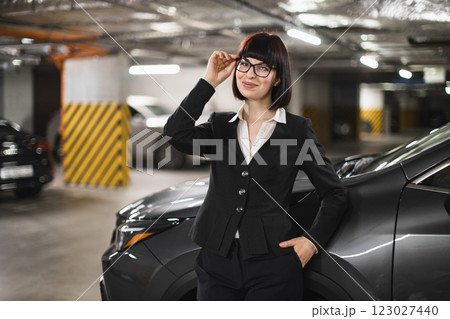 Young adult Caucasian female in business attire standing confidently near car in parking garage, conveying professionalism and empowerment. Young adult Caucasian female in business attire standing confidently near car in parking garage, conveying professionalism and empowerment. 123027440