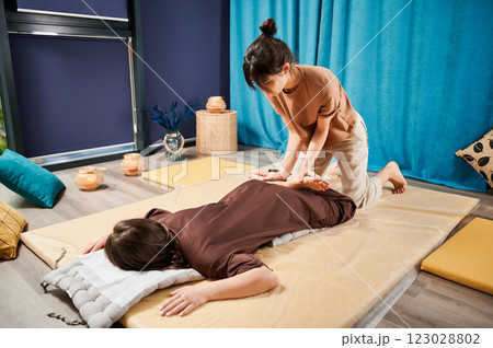 Masseuse making thai yoga massage. Woman therapist doing traditional massage treatment, stretching female patient's body. Client lying on mat, enjoying hand massage in calm atmosphere. Masseuse making thai yoga massage. Woman therapist doing traditional massage treatment, stretching female patient's body. Client lying on mat, enjoying hand massage in calm atmosphere. 123028802