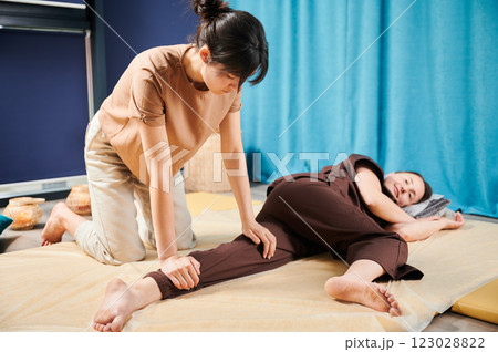 Masseuse making thai yoga massage. Woman therapist doing traditional massage treatment for female patient indoors. Client lying on mat, meditating, enjoying hand massage in calm atmosphere. 123028822