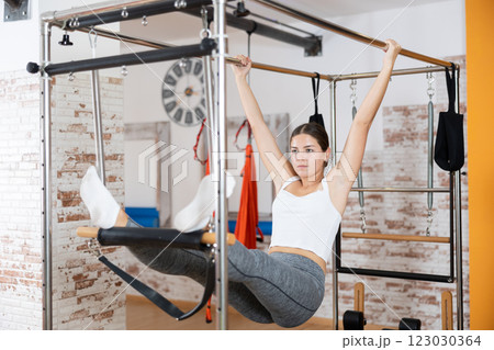 Girl visitor performs exercises in hanging position on combined Pilates trapeze table. 123030364