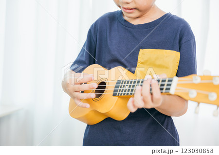A detailed shot of a young boy strumming a yellow ukulele in a cozy living room. His blue shirt contrasts with the soft white curtain backdrop, emphasizing his focus on the music he loves to play. 123030858