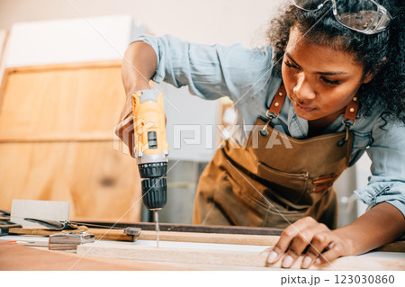 Carpenter woman using a power drill to secure a piece of wood in a workshop, focusing on precision in furniture making. Surrounded by tools and wood pieces, highlighting skill and craftsmanship Carpenter woman using a power drill to secure a piece of wood in a workshop, focusing on precision in furniture making. Surrounded by tools and wood pieces, highlighting skill and craftsmanship 123030860