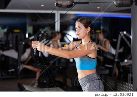 Sportive young girl training her arms on adjustable cable exercise machine in gym Sportive young girl training her arms on adjustable cable exercise machine in gym 123030921
