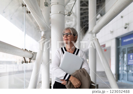Elderly businesswoman stands confidently in a modern office building, holding a laptop and a blazer Elderly businesswoman stands confidently in a modern office building, holding a laptop and a blazer 123032488
