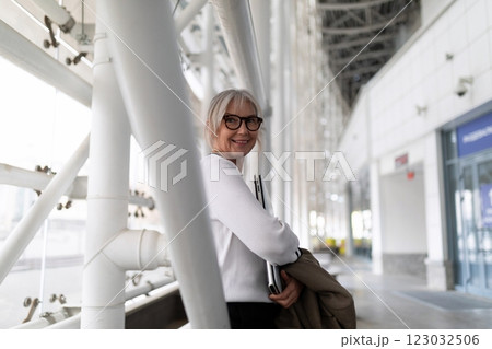 Stylish elderly woman with glasses stands confidently in a modern airport terminal during daytime 123032506