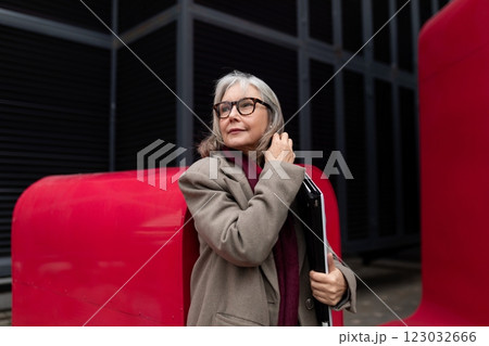 Stylish elderly woman with glasses and a folder poses confidently against vibrant red architectural 123032666