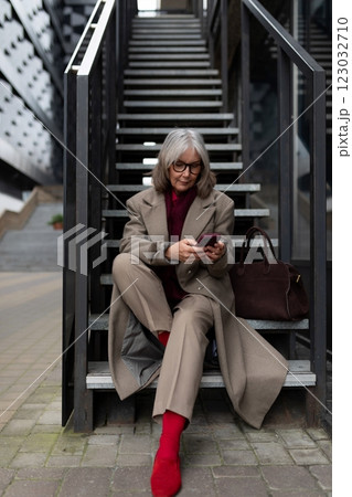 Middle-aged businesswoman in a tailored beige suit sits on stairs checking her phone in an urban Middle-aged businesswoman in a tailored beige suit sits on stairs checking her phone in an urban 123032710