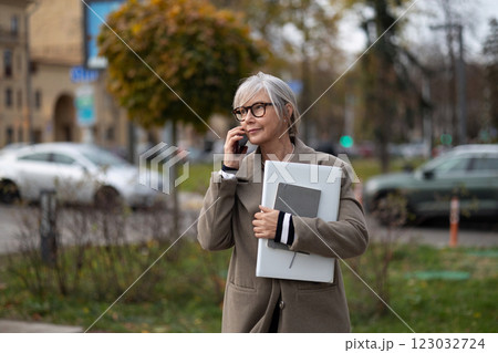 Senior businesswoman walking outside while talking on her phone, holding a laptop and documents on a 123032724