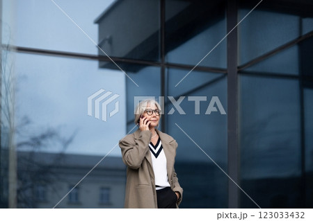 An elderly businesswoman engages in a phone conversation outside a modern office building during the 123033432