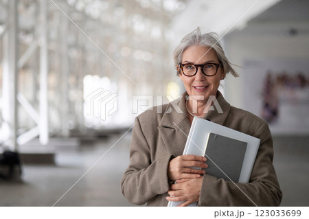 Stylish elderly woman holding a notebook smiles confidently while standing in a modern architectural Stylish elderly woman holding a notebook smiles confidently while standing in a modern architectural 123033699