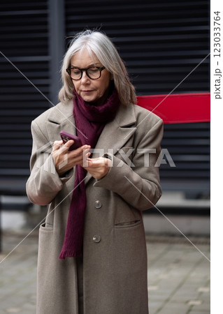 An older woman dressed warmly checks her phone while standing outdoors on a chilly day in an urban 123033764