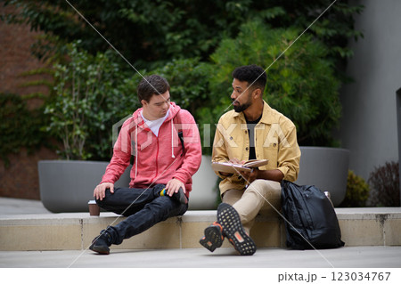 Happy young man with Down syndrome and mentoring friend sitting and talking outdoors, spending time together. 123034767