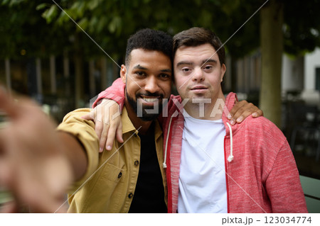 Young man with Down syndrome and his volunteer buddy taking selfie with smatphone, sitting outdoors. 123034774