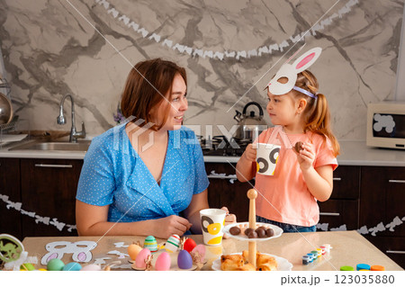 The Easter Spring festival. Mid shot of Caucasian mother and daughter drink tea and eat festive treats together, sitting in a decorated kitchen. Pre-school children's education 123035880