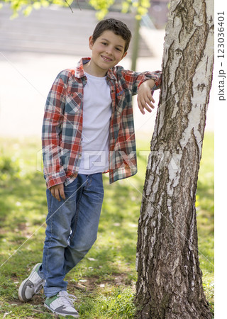 Smiling boy in plaid shirt leaning against a tree in a park. 123036401