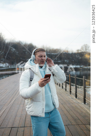 Smiling young man is walking near a lake on the forest, wearing headphones and using a mobile phone. Smiling young man is walking near a lake on the forest, wearing headphones and using a mobile phone. 123036425